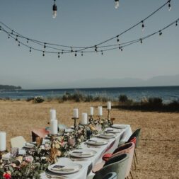 Long table with white tablecloth and place settings with candles and flowers set up at the beach with string lights hanging overhead.