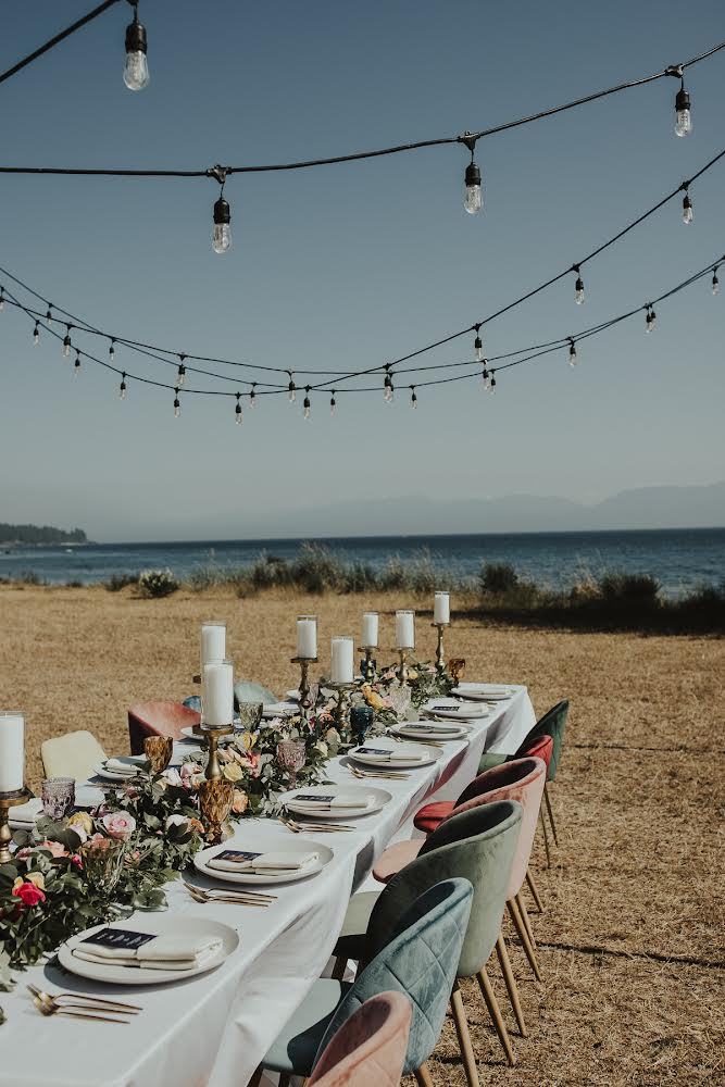 Long table with white tablecloth and place settings with candles and flowers set up at the beach with string lights hanging overhead.